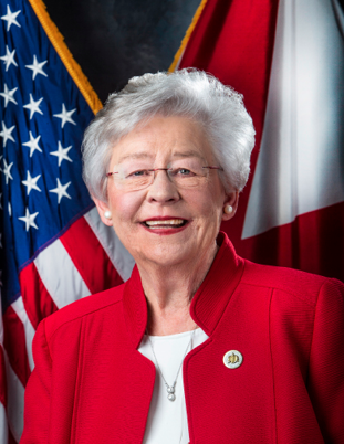 Official portrait of Alabama Governor standing in front of the U.S. and Alabama state flags, wearing a red blazer and smiling.