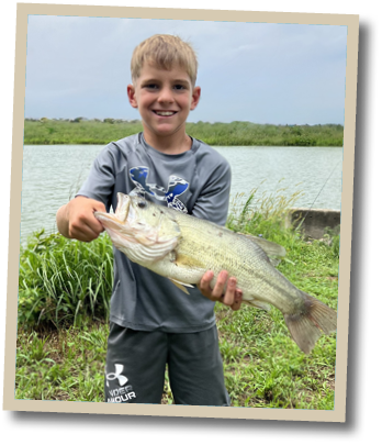 Young angler smiling while holding a largemouth bass near a grassy freshwater pond in Delaware.