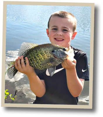 Young angler smiling while holding a black crappie near the edge of a freshwater lake in Delaware.