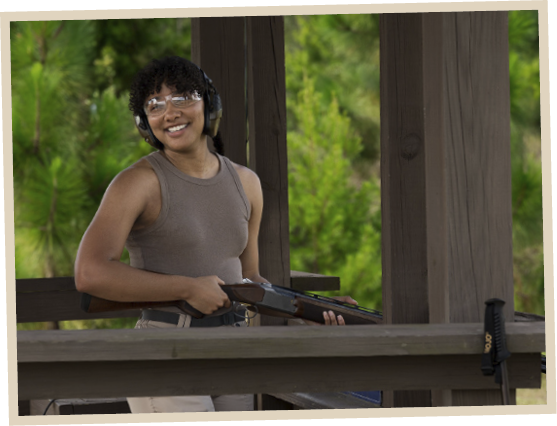 Smiling woman wearing safety glasses and earmuffs holding a shotgun at an outdoor shooting range surrounded by trees.