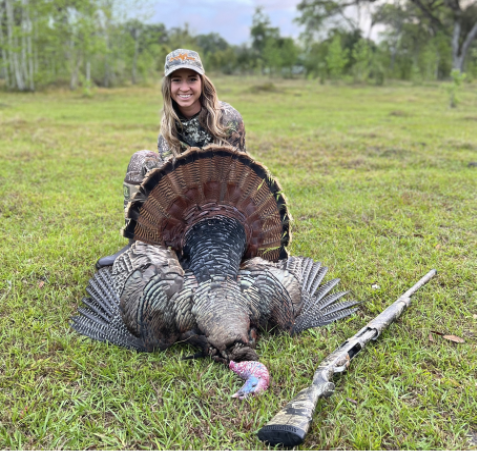Smiling female hunter in camouflage kneels behind a harvested wild turkey displayed on grass, with a shotgun lying beside them.