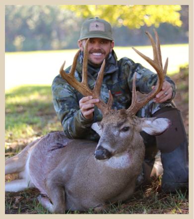 Smiling hunter in camouflage gear poses with a large white-tailed buck during deer season in a grassy, wooded area.