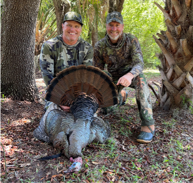 Two smiling hunters in camouflage pose with a harvested wild turkey in a Florida woodland setting.