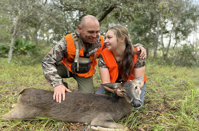 Father and daughter in hunter orange vests smiling together after a successful deer hunt in Florida, kneeling beside a harvested deer in a wooded area.