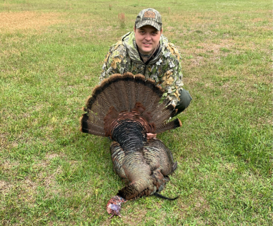 Hunter in camouflage kneeling on grass behind a harvested wild turkey with its tail feathers fanned out.