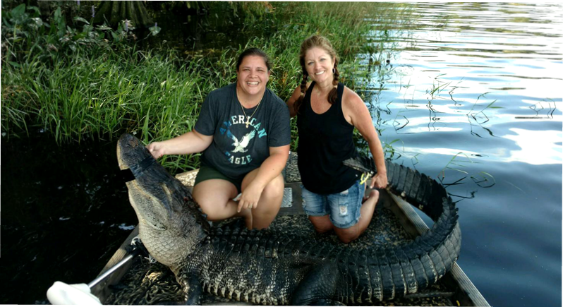 Two women sitting in a boat with a harvested American alligator after a successful Florida alligator hunt. The large gator is curled around them as they smile for the photo near the water's edge.