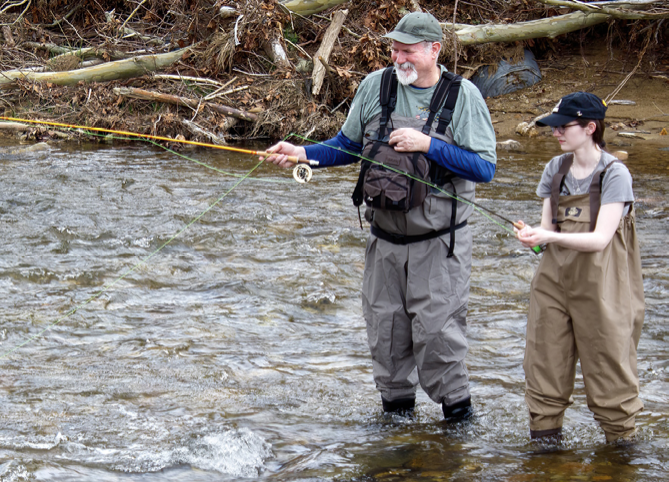 An older man and a young woman wearing waders fly fishing together in a shallow stream surrounded by fallen branches and rocks.