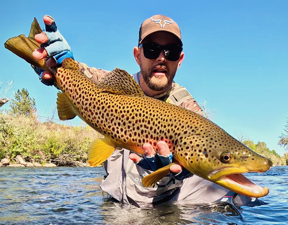 Angler standing in a river holds a large brown trout with spotted markings, showcasing a successful catch during fishing in Nevada waters.