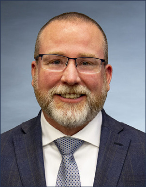 Portrait of a smiling man with glasses, a beard, and a navy suit with a light blue tie, posed in front of a soft blue background.