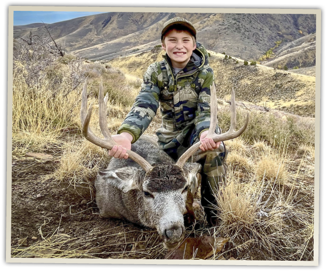 Young Nevada hunter smiling while holding the antlers of a harvested mule deer buck in mountainous Nevada hunting terrain.