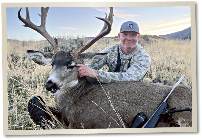Nevada hunter posing with a harvested mule deer buck with large antlers in a sagebrush field, with a rifle resting beside the deer during a Nevada big game hunt.