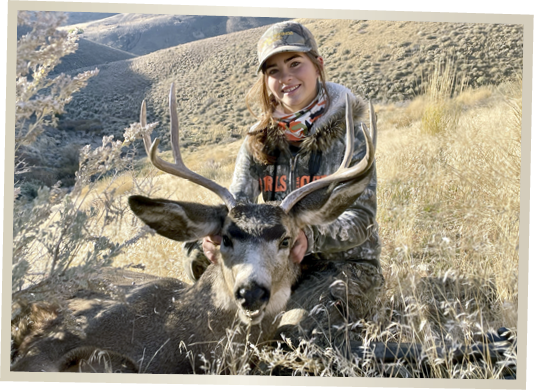 Young hunter posing with a harvested mule deer buck with antlers in Nevada sagebrush hills during a big game hunting trip.