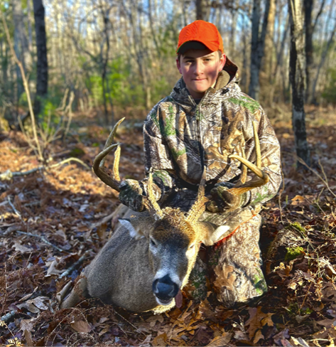 Young hunter wearing camouflage and an orange hat kneels beside a harvested white-tailed deer in a wooded area during Rhode Island’s deer hunting season.