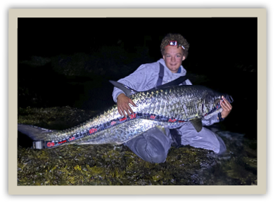 Thomas Czernik kneels on a rocky shoreline at night, holding a massive 60+ inch tarpon caught while shore fishing on Aquidneck Island.