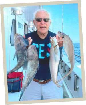Orlando Savastano smiles while holding a mixed catch of black sea bass and hake on the deck of the Gail Frances fishing boat with the ocean in the background.