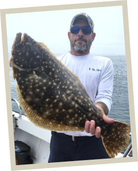 Kevin Rahilly stands on a boat holding a large summer flounder, also known as a "doormat," with calm ocean waters in the background.