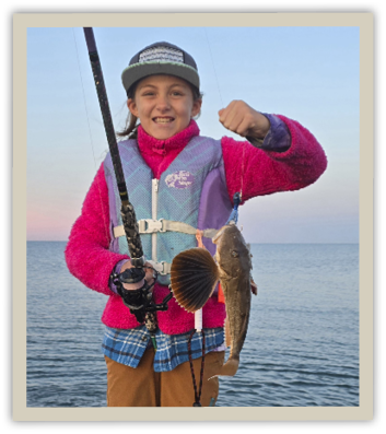 Young angler Rosie Clifton proudly holds up a striped searobin she caught using a bucktail jig she tied herself, standing by the ocean at sunrise.